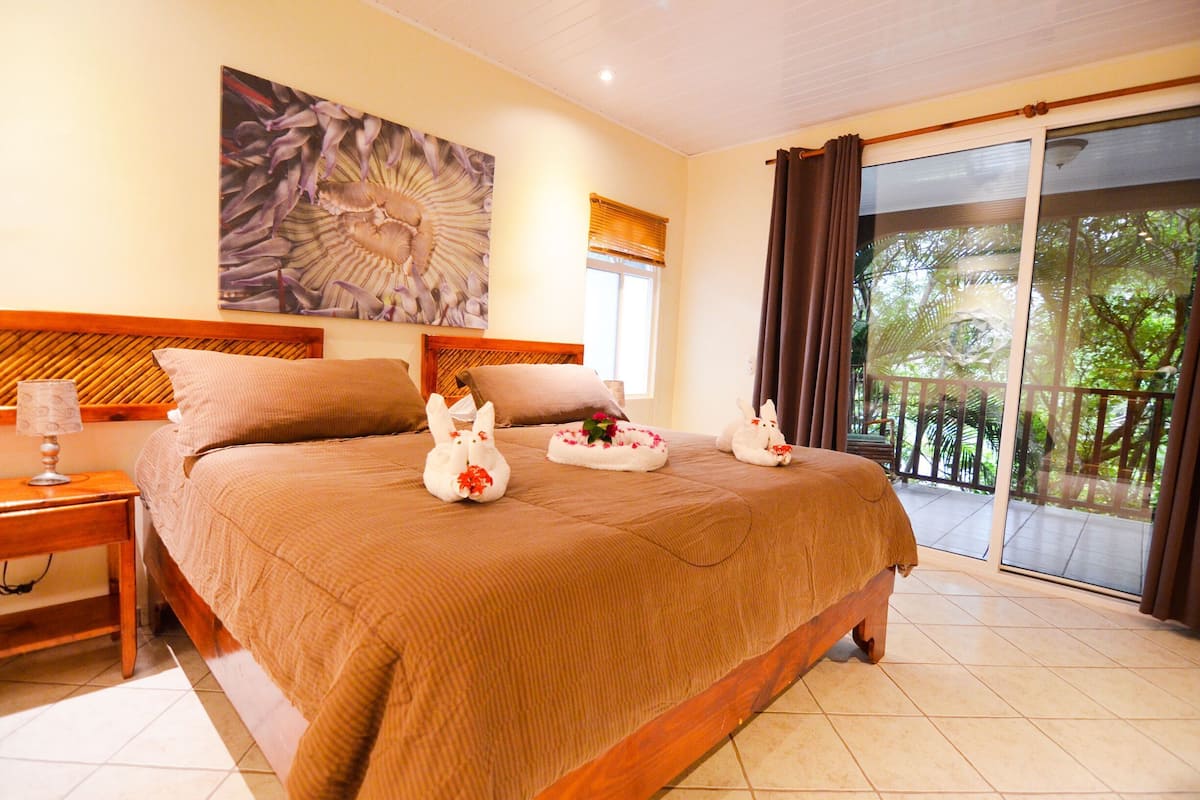 Habitación de hotel con una cama grande, cabecera de madera, mesita de noche, lámpara y vistas al exterior a través de una puerta corrediza de vidrio.
