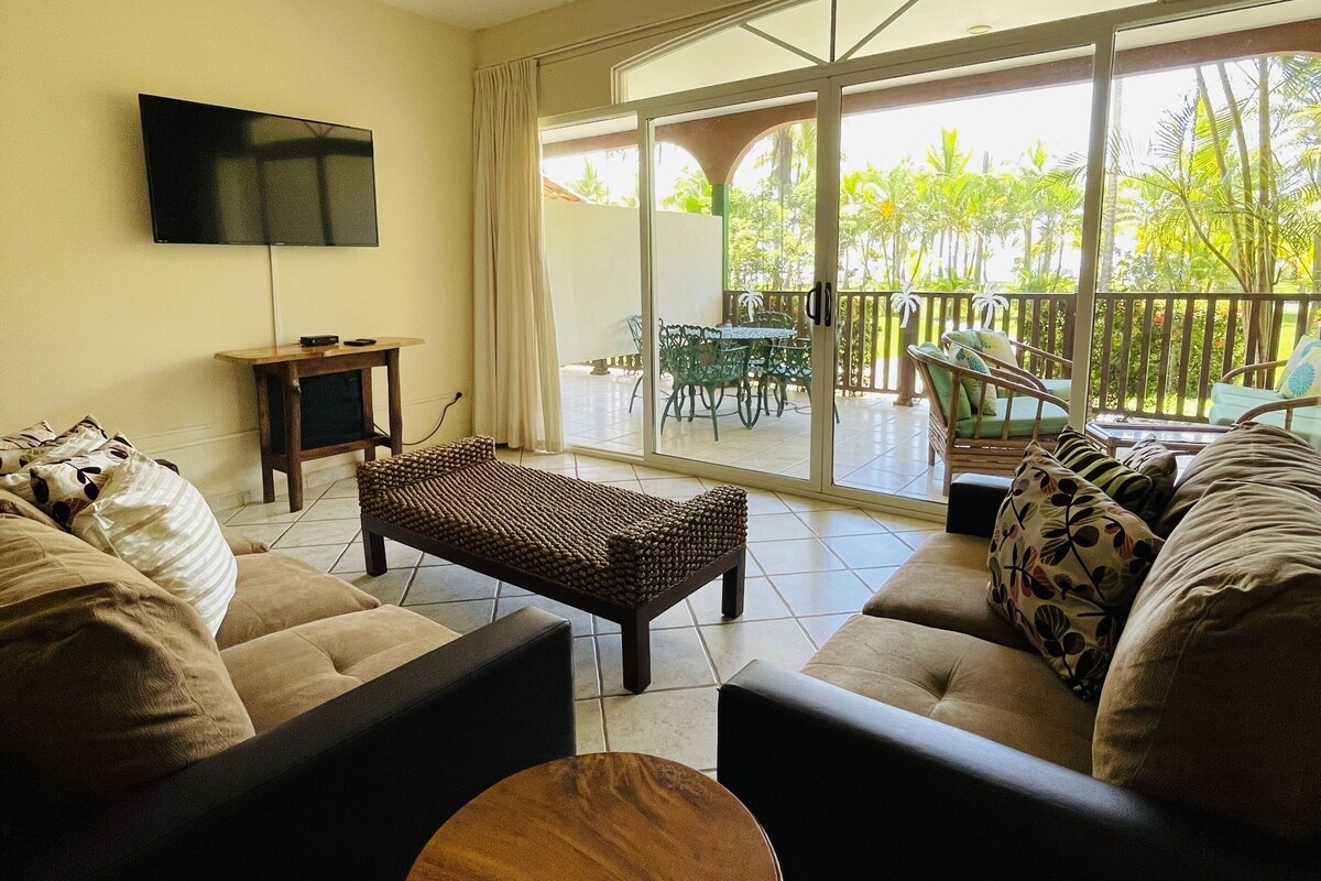 A living room with a sofa, a coffee table, a TV, and a balcony with outdoor furniture.