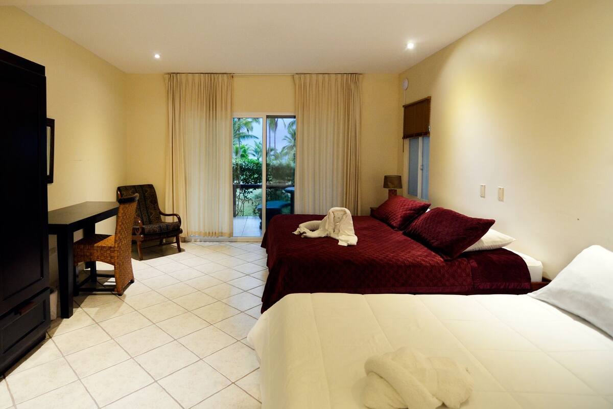 Habitación de hotel con una cama, una silla, un escritorio y vista a palmeras a través de la ventana.