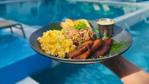 Eine Person hält einen Teller mit Essen neben einem Pool, umgeben von viel Grün und blauem Himmel.