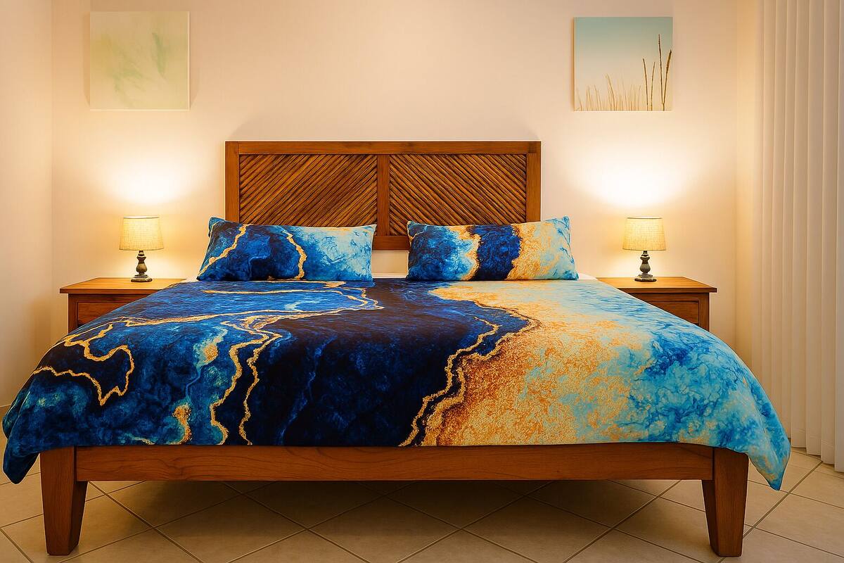 Una cama con una colcha de diseño azul y dorado, cabecera de madera, dos lámparas de noche y una mesita de noche.