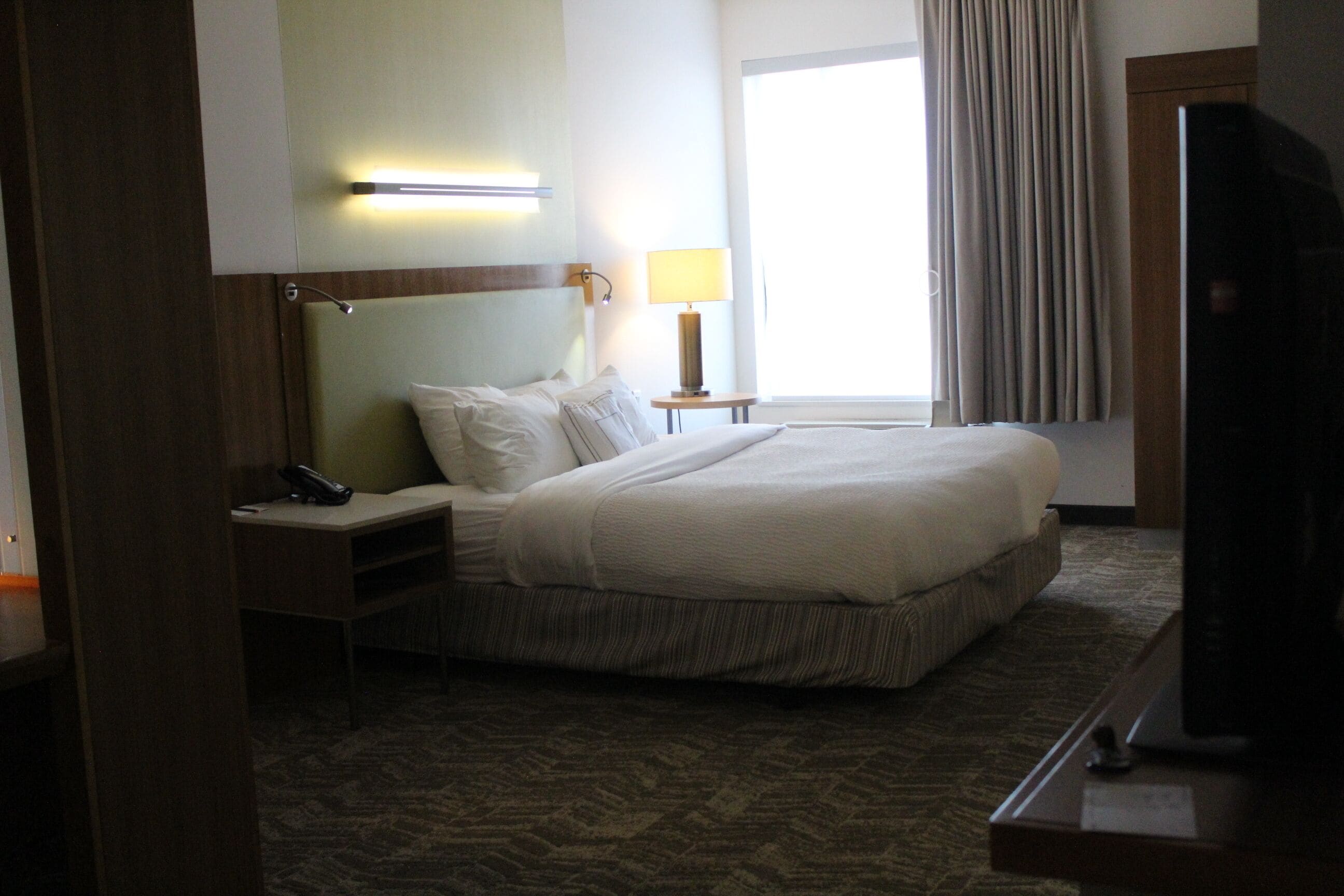 A hotel room with a bed, bedside table, lamp, TV, and a window with curtains.