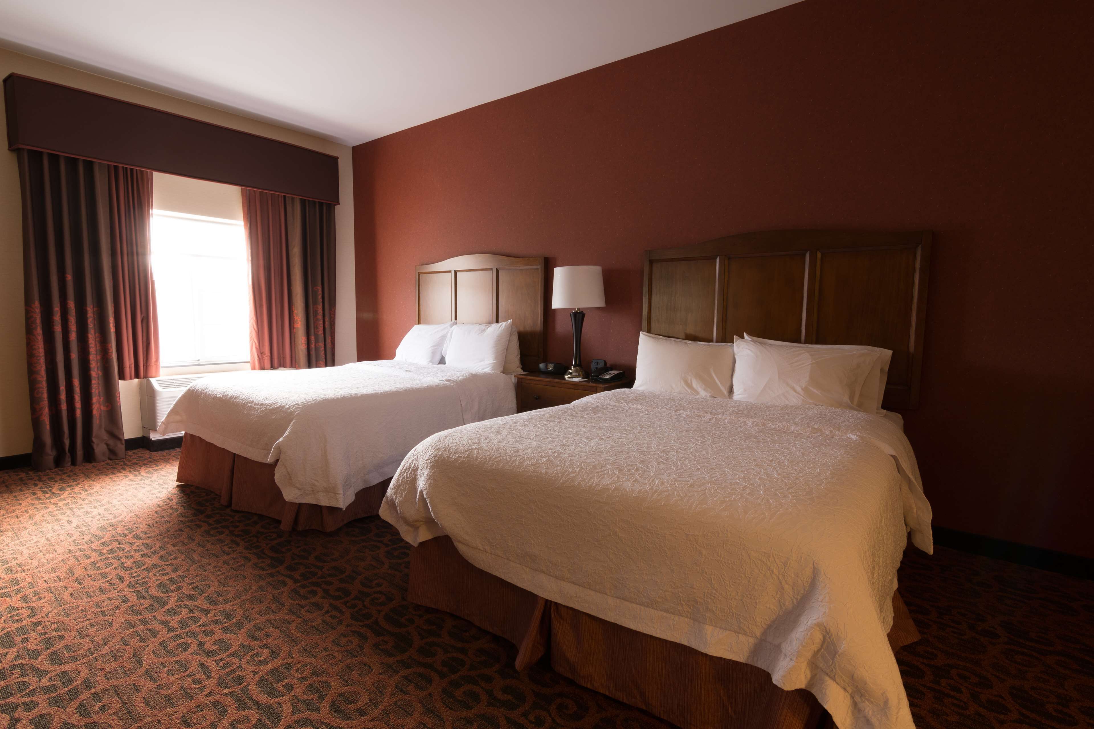 A hotel room with two beds, a nightstand with a lamp, and a window with curtains.