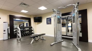 Academia com equipamentos de musculação, um banco e uma televisão fixada na parede.