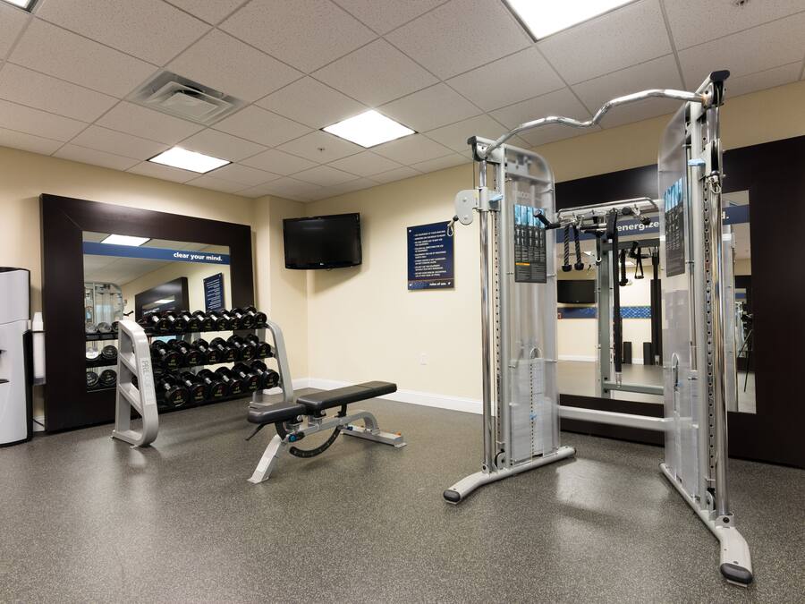Academia com equipamentos de musculação, um banco e uma televisão fixada na parede.