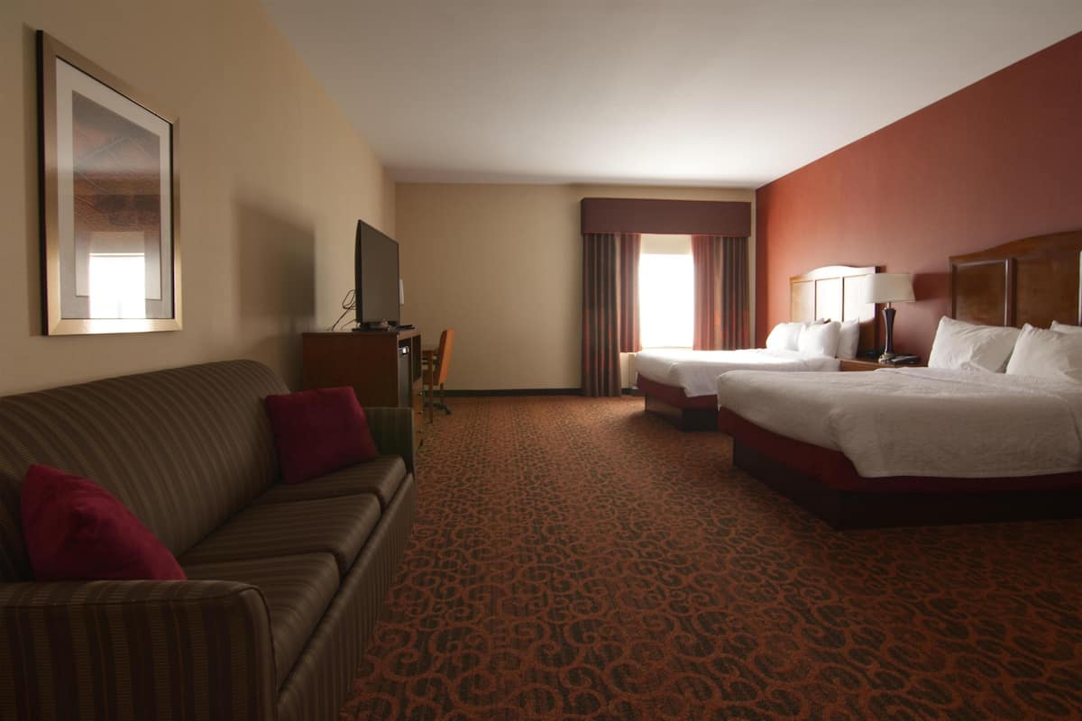A hotel room with a sofa, two beds, a TV, and a large window with curtains.