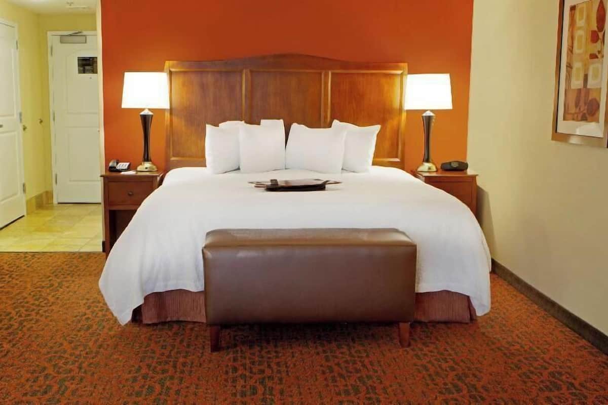 A hotel room with a large bed, two bedside tables, a wooden headboard, a bench at the foot of the bed, and a door in the background.