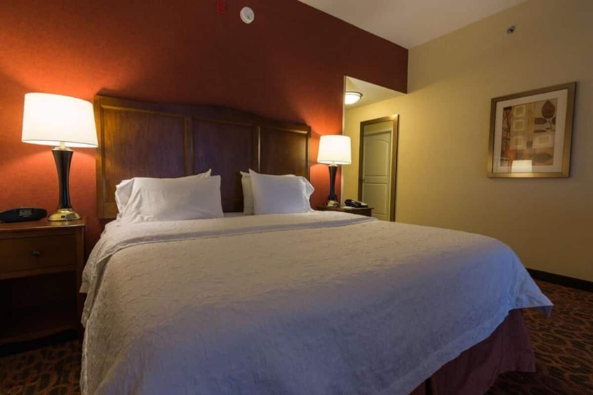 A hotel room with a large bed, two bedside tables with lamps, a nightstand, a door, and a framed picture on the wall.
