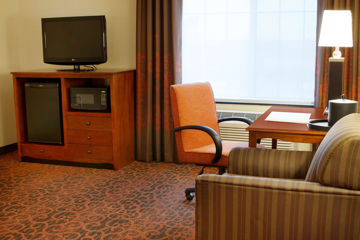 A hotel room with a television on a wooden cabinet, a microwave, a chair, a table, and a lamp.