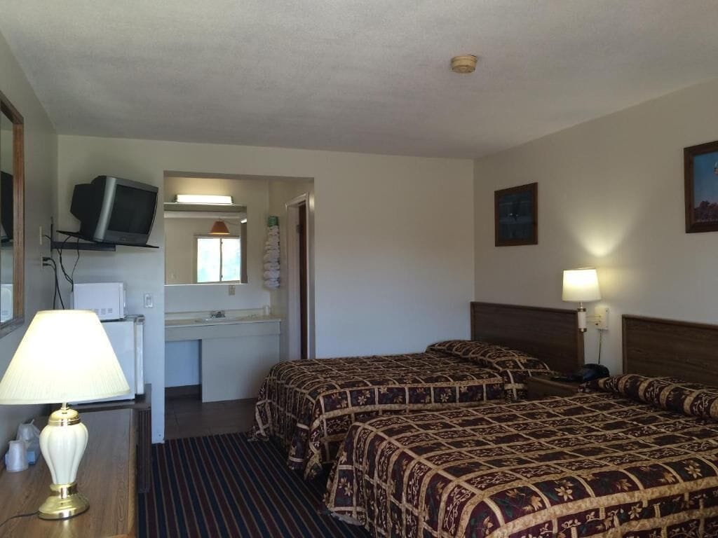 A hotel room with two beds, a TV on a shelf, a desk with a lamp, and a bathroom visible through an open door.