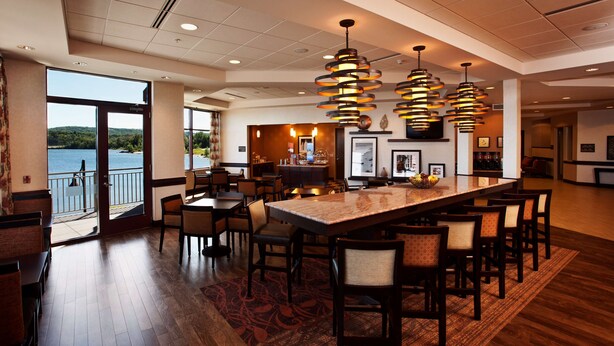A spacious restaurant with a large central table, multiple chairs, and a view of a lake. - Hampton Inn Marquette/Waterfront (Marquette)