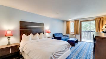 Une chambre d’hôtel avec un grand lit, deux lampes de chevet, un fauteuil bleu, un bureau et une vue sur l’extérieur grâce à une porte-fenêtre coulissante.