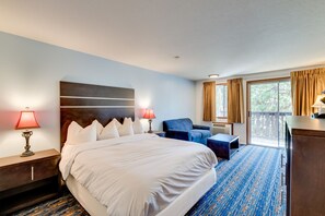 A hotel room with a large bed, two bedside lamps, a blue armchair, a desk, and a view of the outdoors through a sliding glass door.