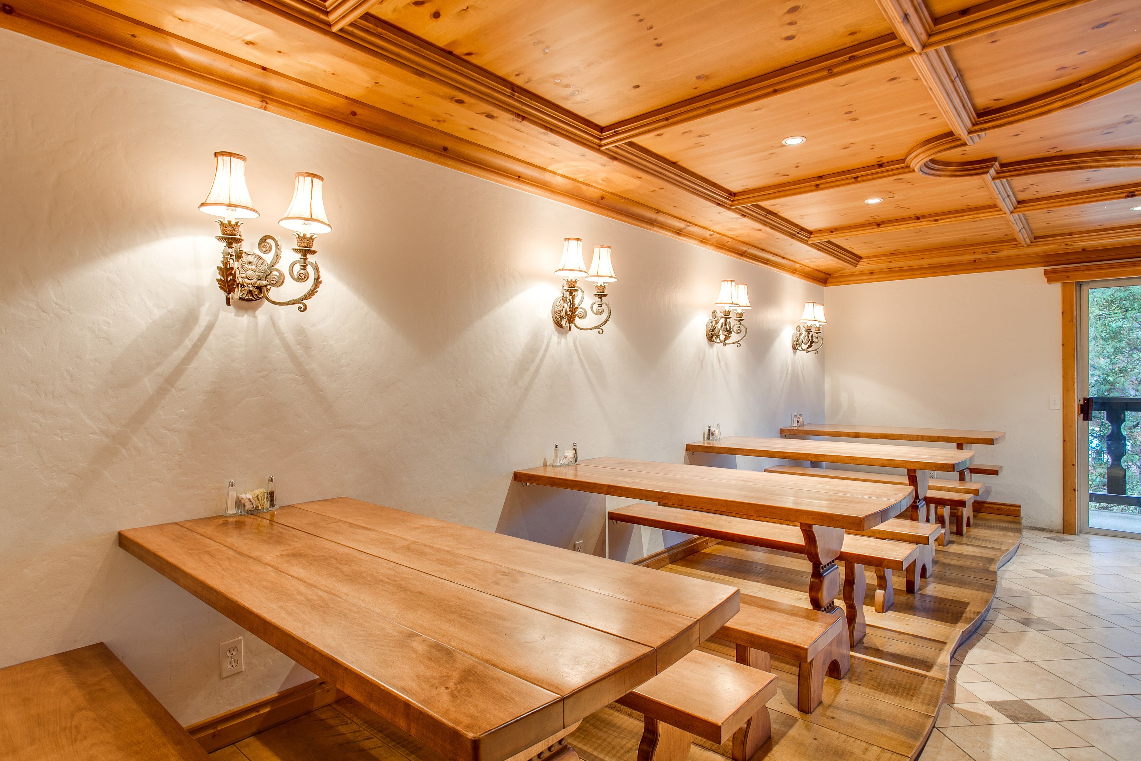 Un comedor con mesas y bancos de madera, lámparas fijadas a la pared y vistas al exterior a través de un ventanal grande.