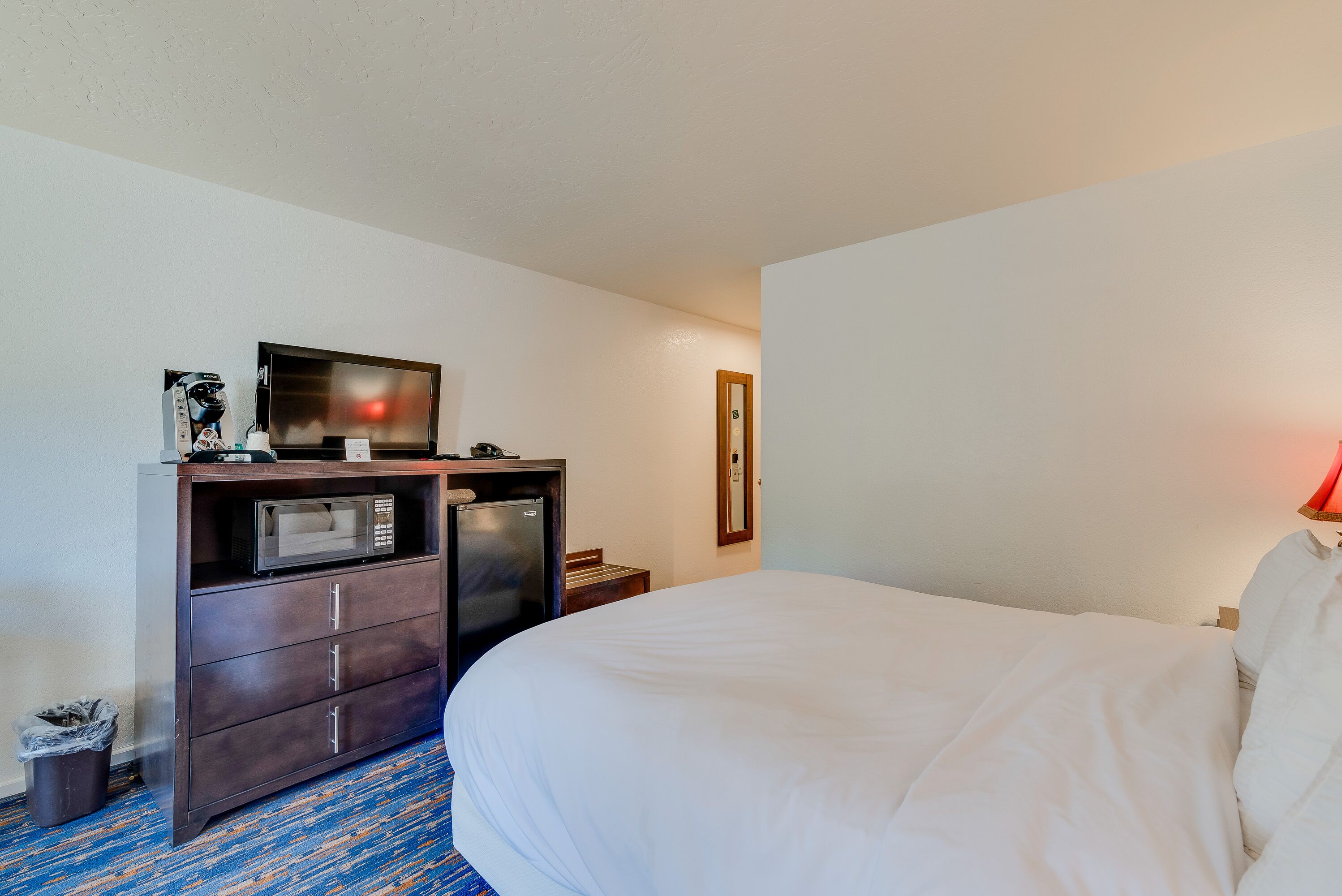 A hotel room with a bed, a desk with a microwave, a trash bin, and a mirror on the wall.