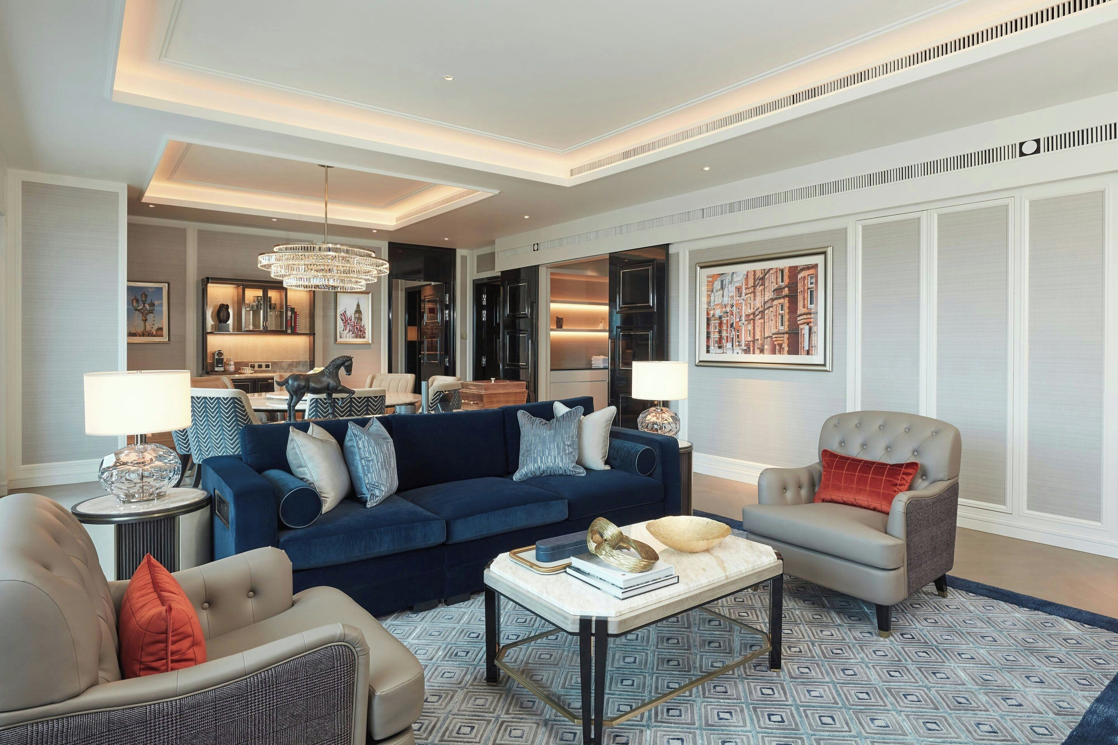 A spacious living room with a dark blue sofa, a grey armchair, and a coffee table. There is a chandelier hanging from the ceiling and a framed cityscape painting on the wall.
