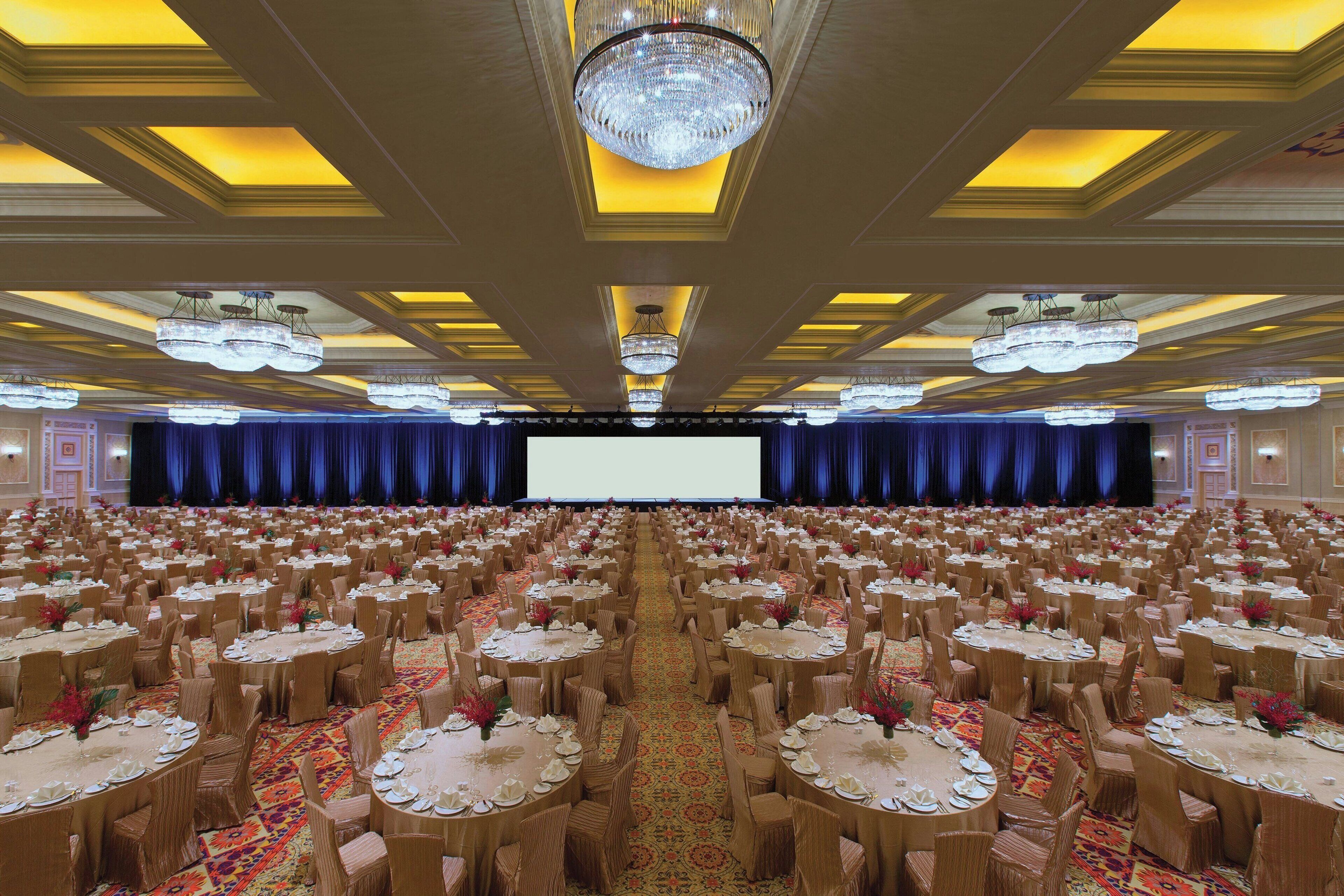 A banquet hall with round tables, chairs, a stage, and a large screen.