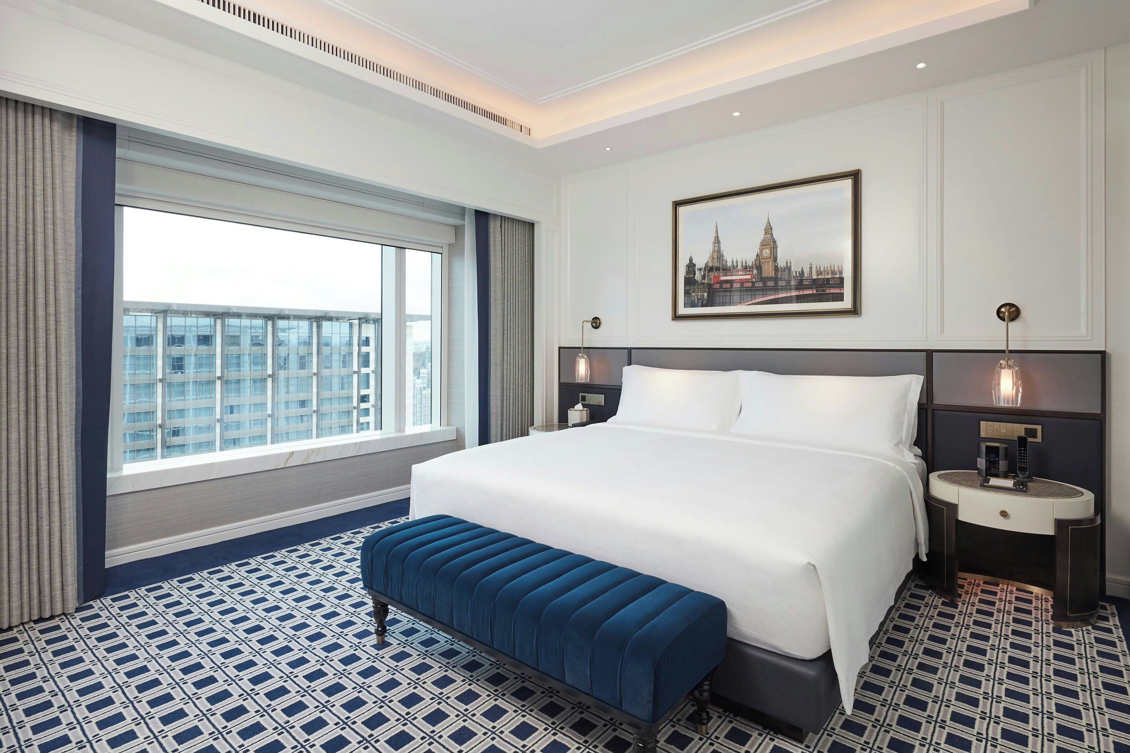 Une chambre d’hôtel avec un grand lit, une table de chevet, une commode, une fenêtre avec des rideaux, une moquette bleue à motifs et une vue sur la ville.