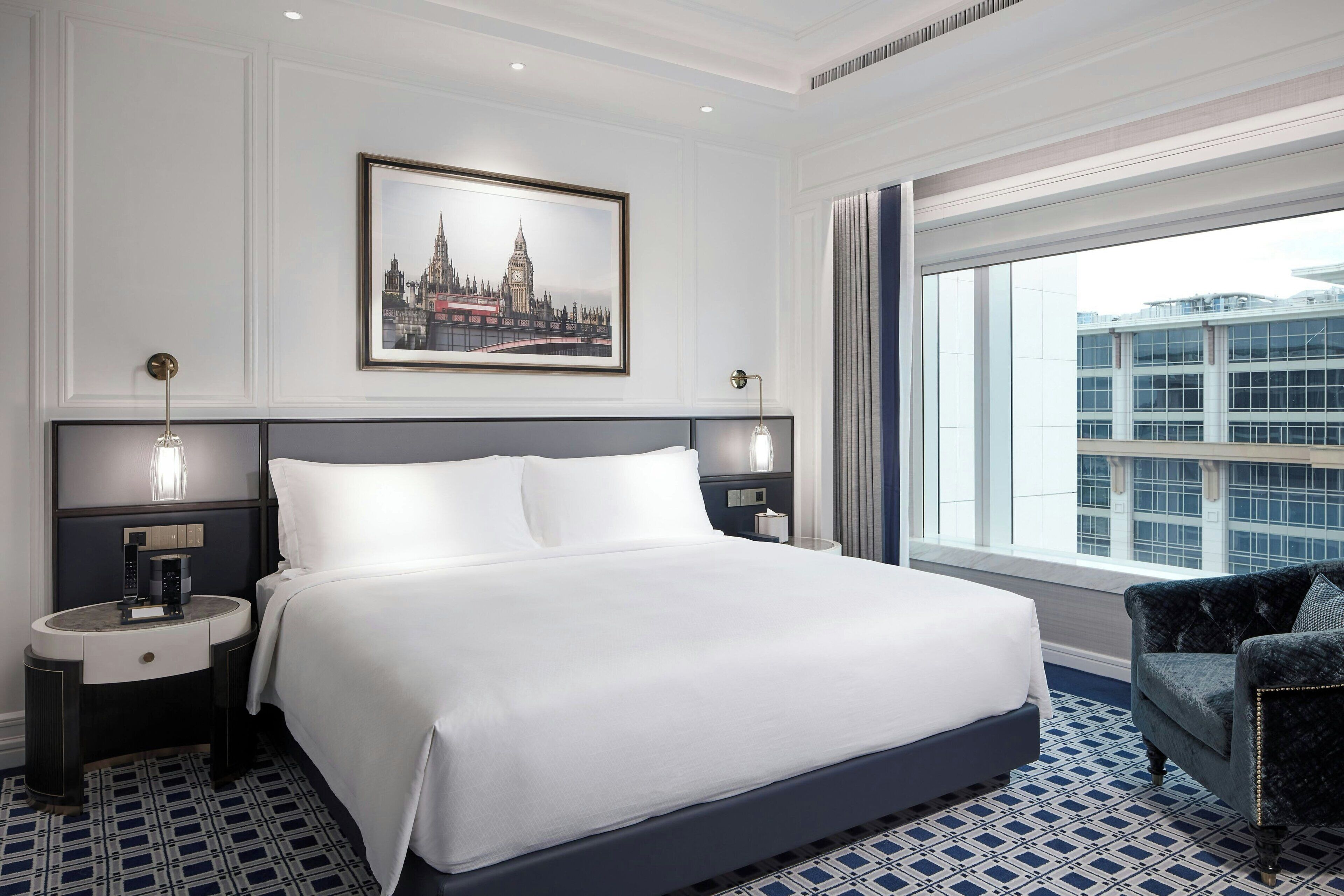 Une chambre d’hôtel avec un grand lit, une table de chevet, une chaise, un tableau représentant une vue sur la ville, et une fenêtre donnant sur la ville.