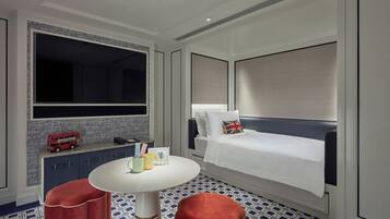 Quarto de hotel com uma cama grande, uma mesa de cabeceira, uma mesa redonda e um banquinho estofado vermelho.