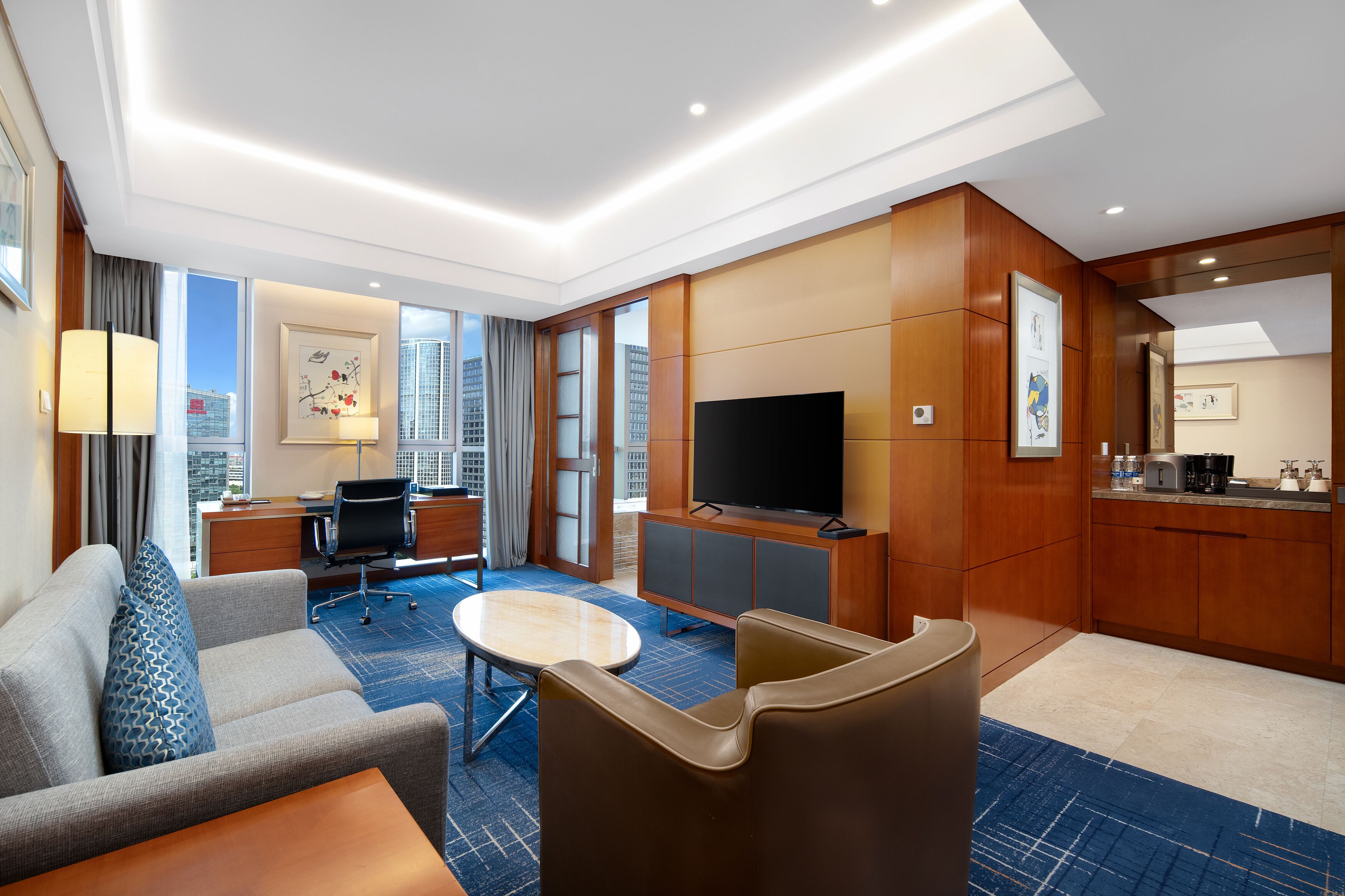 A hotel room with a sofa, armchair, desk, TV, and a city view.