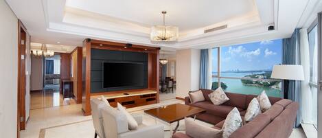 A spacious living room with a large flat-screen TV, comfortable seating, and a view of a body of water through large windows.