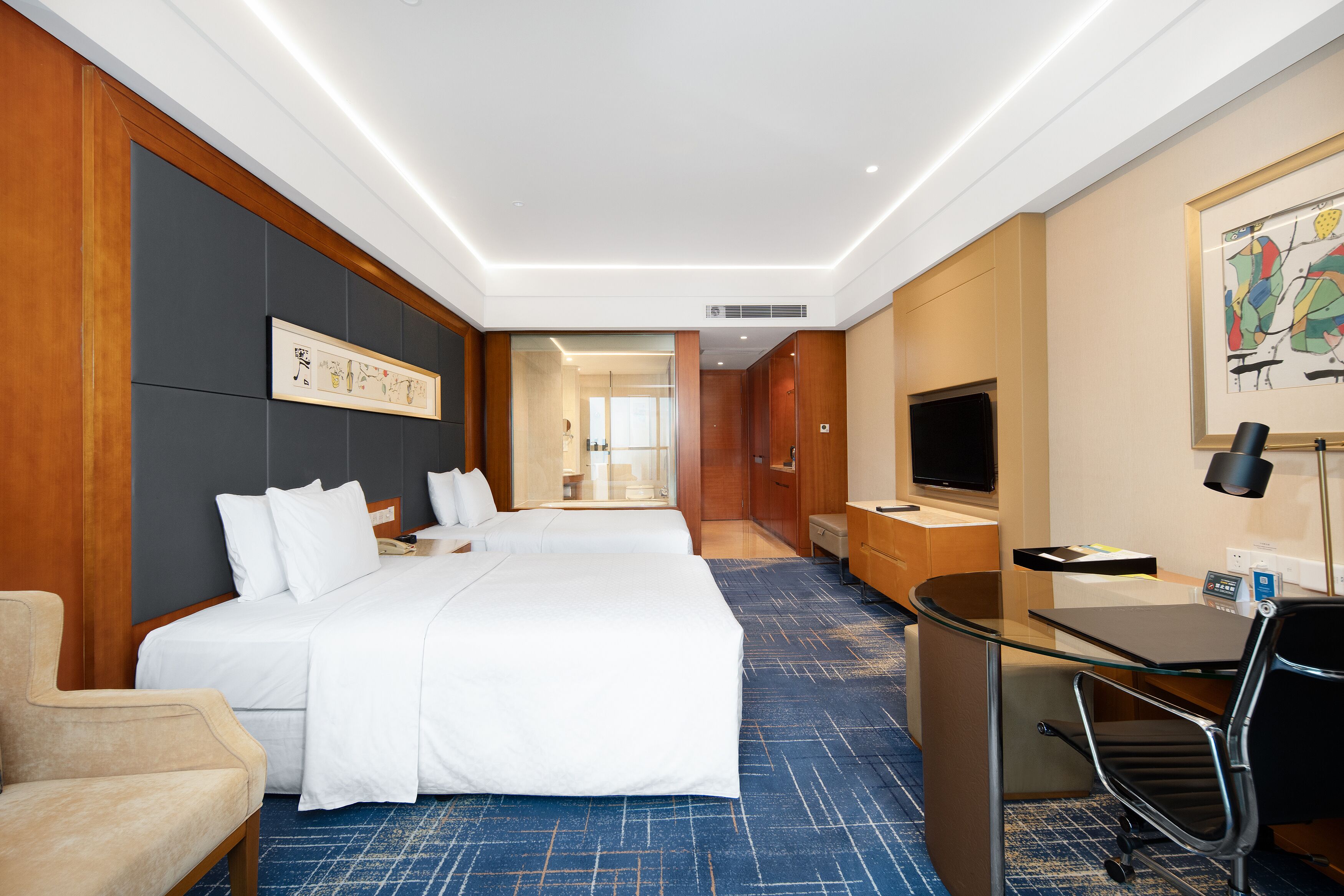 A hotel room with two beds, a desk, a chair, a TV, and a painting on the wall.