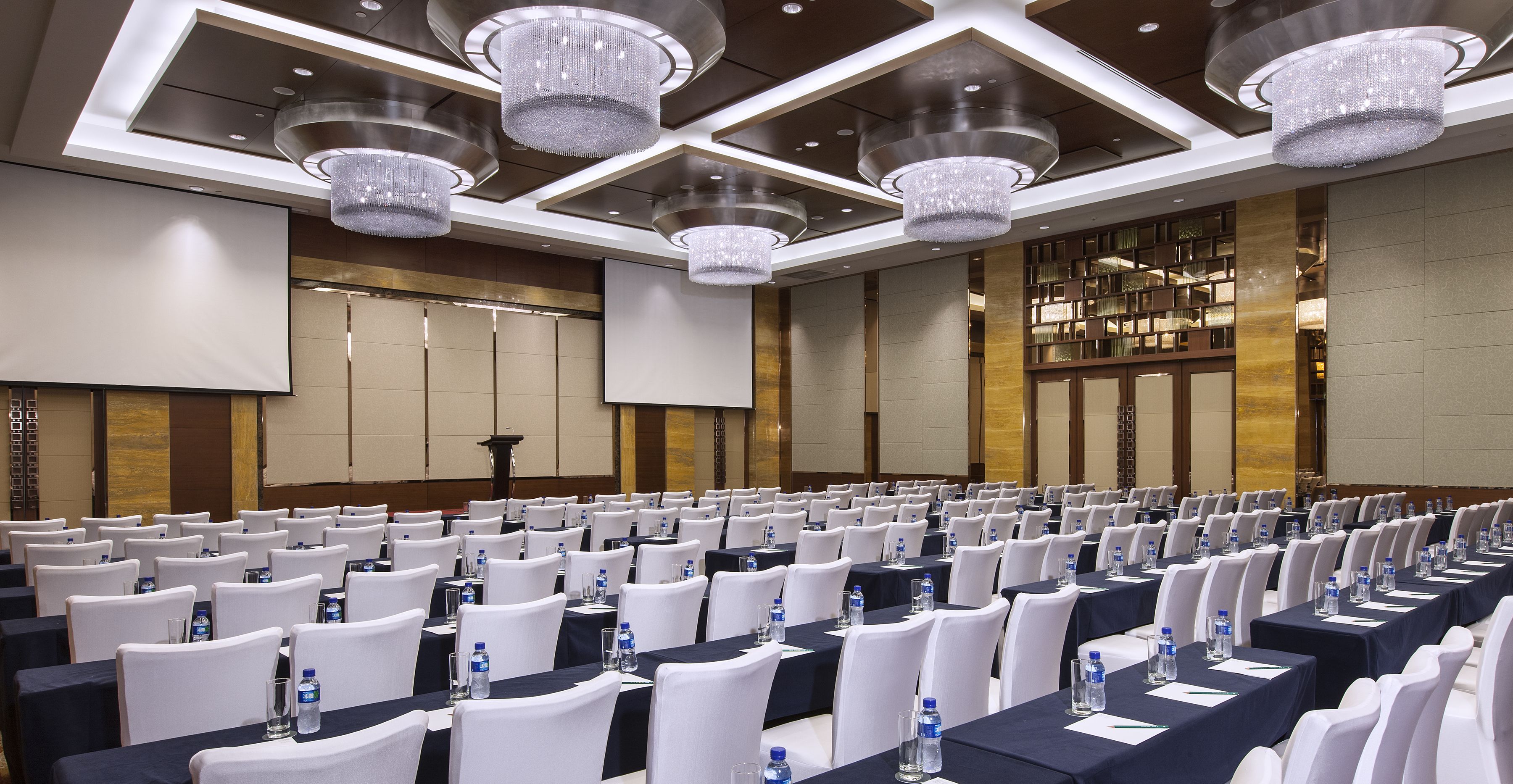 Un salón de conferencias con filas de mesas, sillas blancas y botellas de agua.