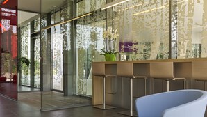 A modern reception area with a glass wall, a reception desk, a blue chair, and a white table. - ibis Leuven Heverlee (Leuven)