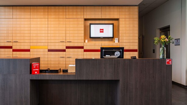 A hotel reception desk with a mounted television, a digital scale, and a sign indicating no smoking. - ibis Leuven Heverlee (Leuven)