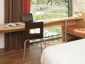 A hotel room with a wooden desk, a laptop, a chair, a bed, and a window with a view of greenery and buildings. - ibis Leuven Heverlee (Leuven)