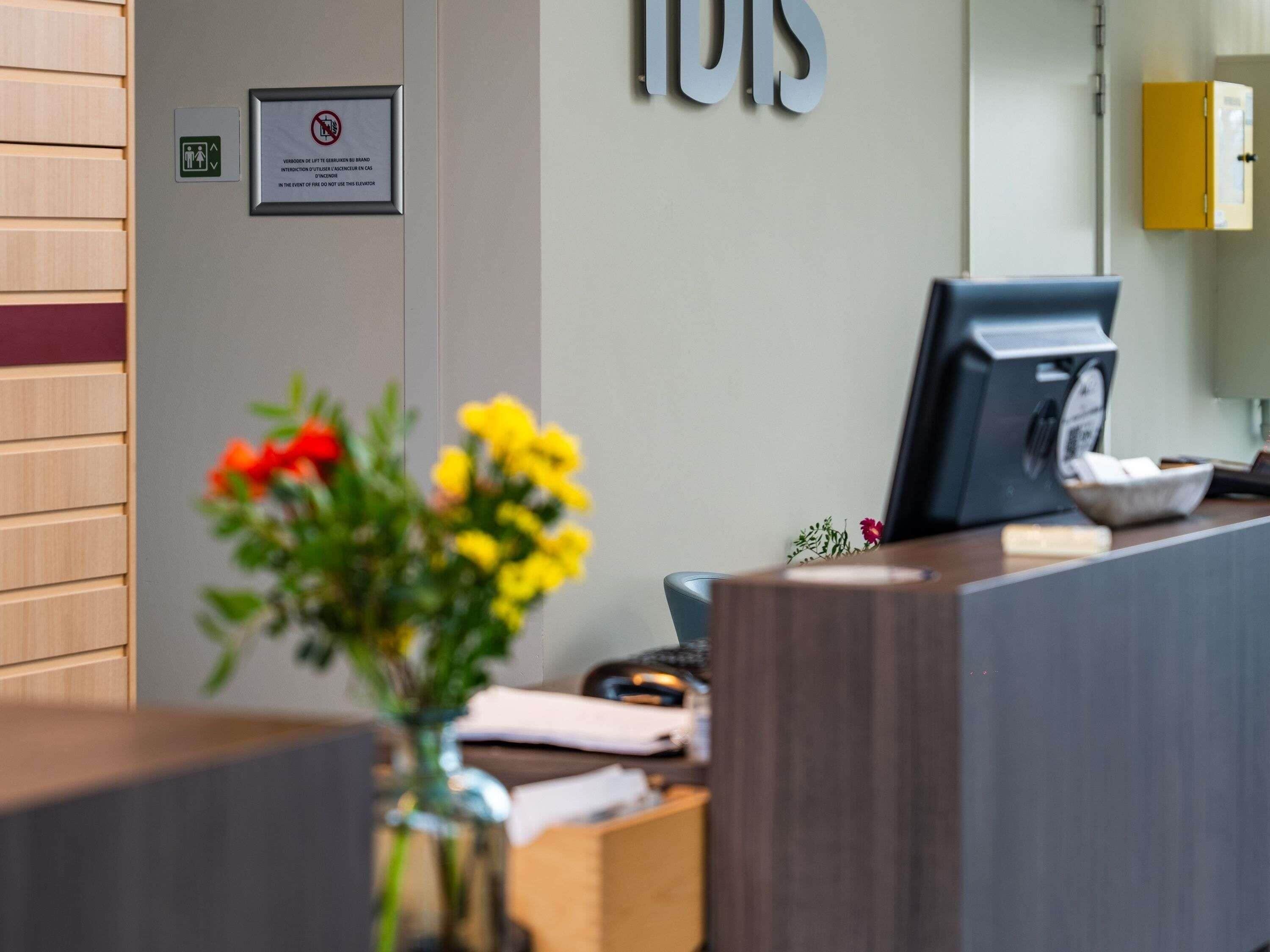 A reception desk with a framed sign, a computer monitor, and a vase with flowers.