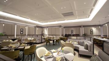 A spacious hotel dining area with multiple tables and chairs, a central seating area, and a modern ceiling design.