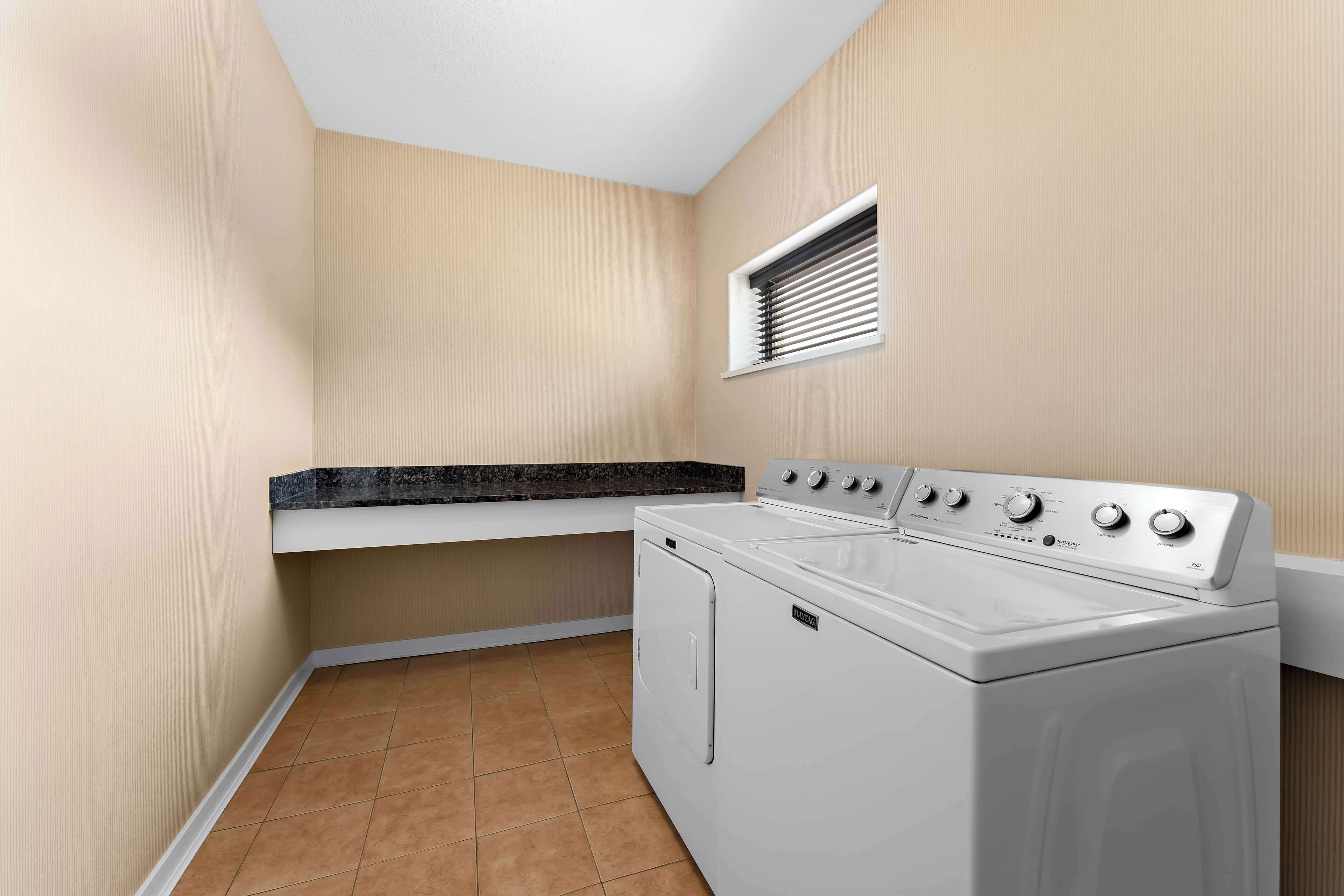 A laundry room with a washing machine and dryer, a small window with blinds, and a countertop.