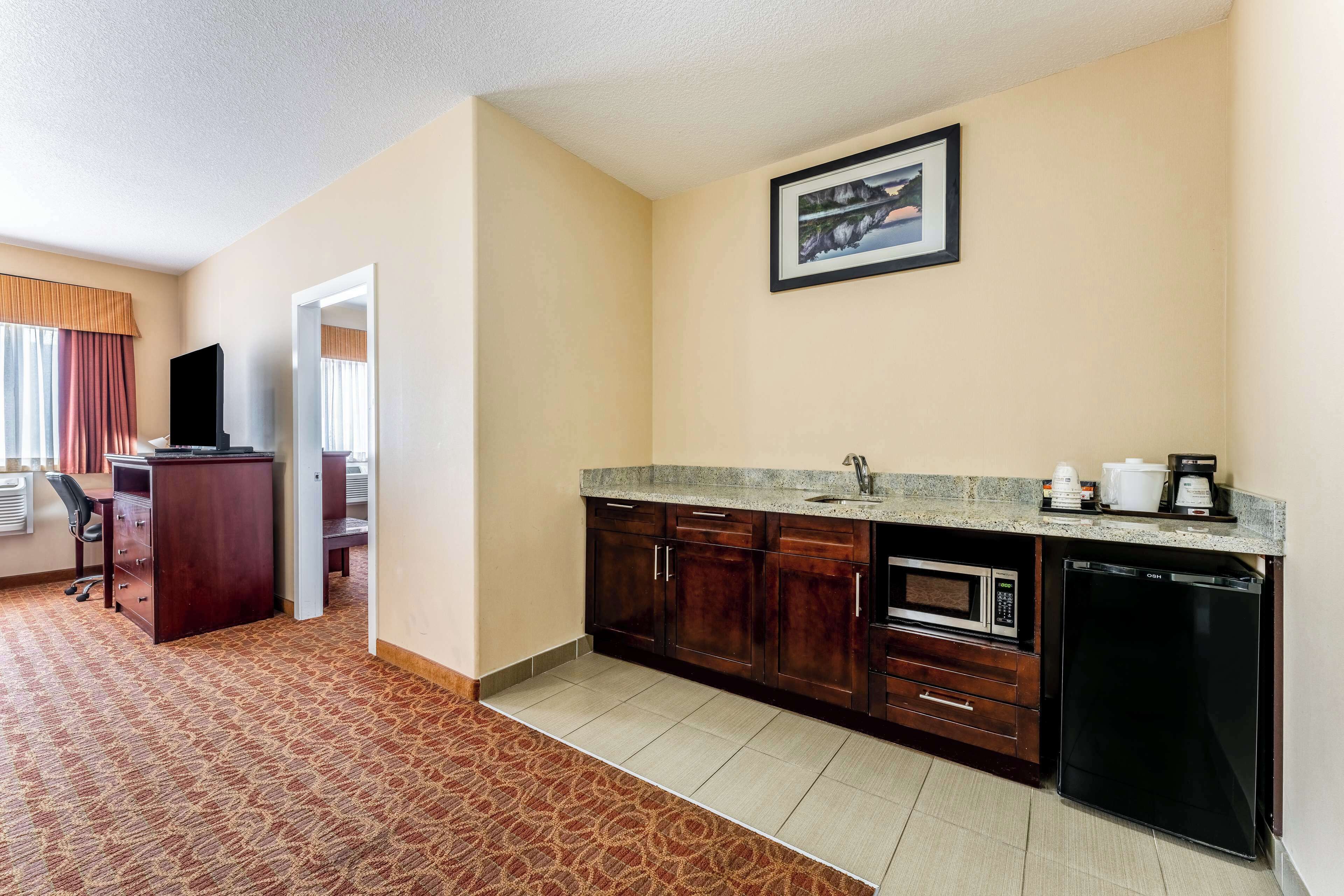 A compact hotel room with a kitchenette, a bed, a desk, and a window with curtains.