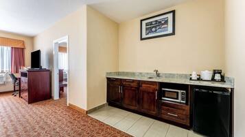 A compact hotel room with a kitchenette, a bed, a desk, and a window with curtains.