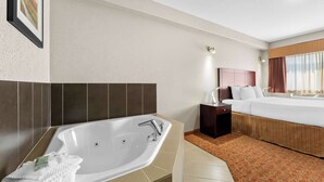Habitación de hotel con una bañera grande, una cama con cabecera, una mesita de noche y un ventanal con cortinas.