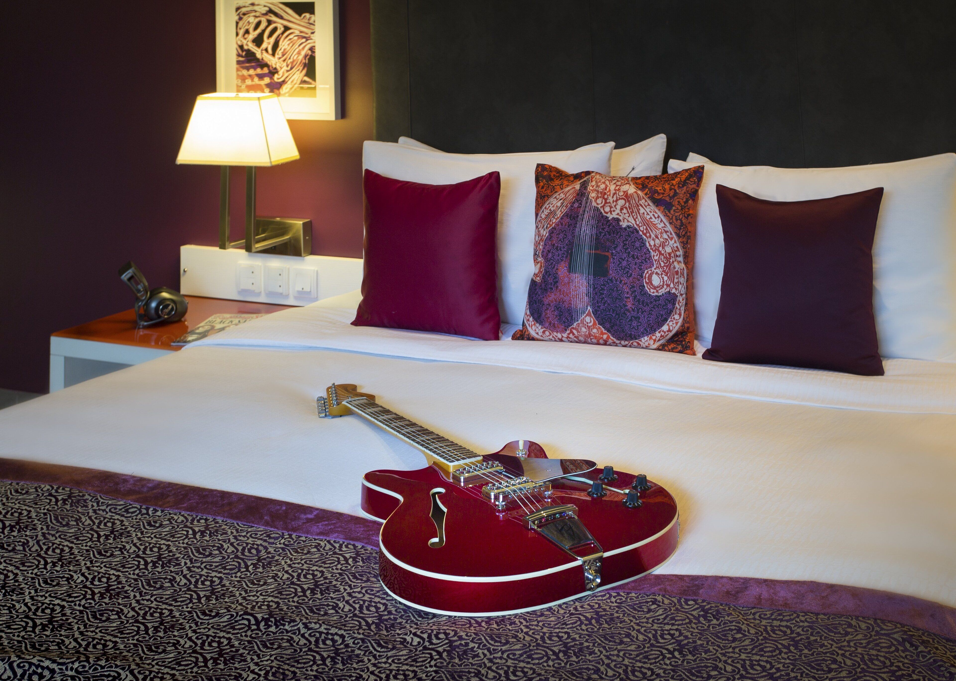 A hotel room with a neatly made bed, a red guitar on the bed, and a lamp on the nightstand.