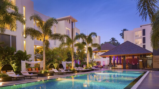 A swimming pool with lounge chairs and palm trees, surrounded by modern buildings with balconies and a bar area. - Hard Rock Hotel Goa Calangute (Calangute)