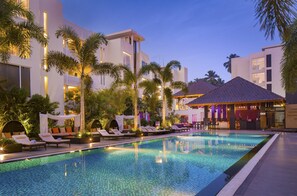 Una piscina con sillas de descanso y palmeras, rodeada de edificios modernos con balcones y una zona de bar.