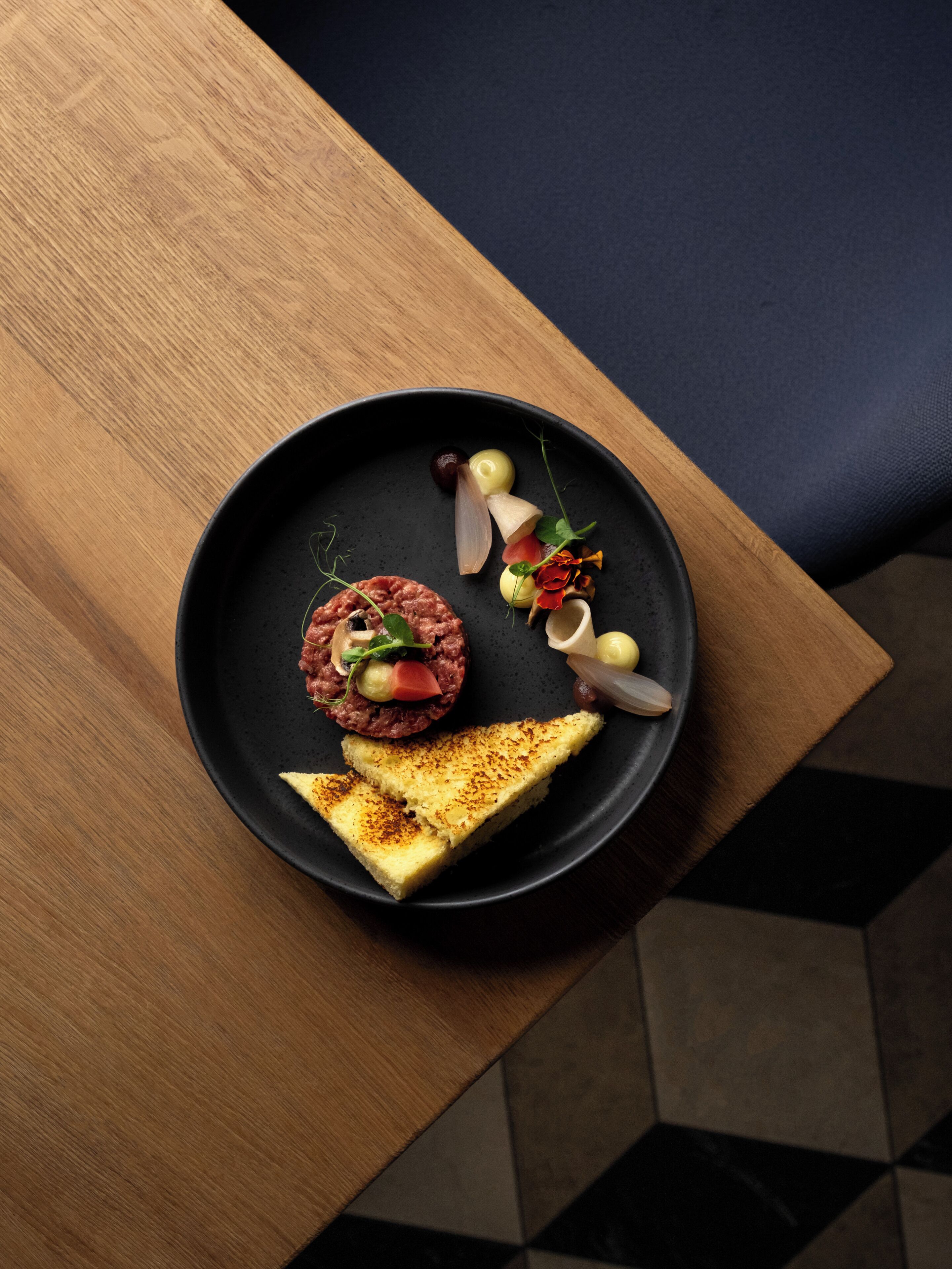 A black plate with a steak tartare, pickles, and a slice of bread on a wooden table.