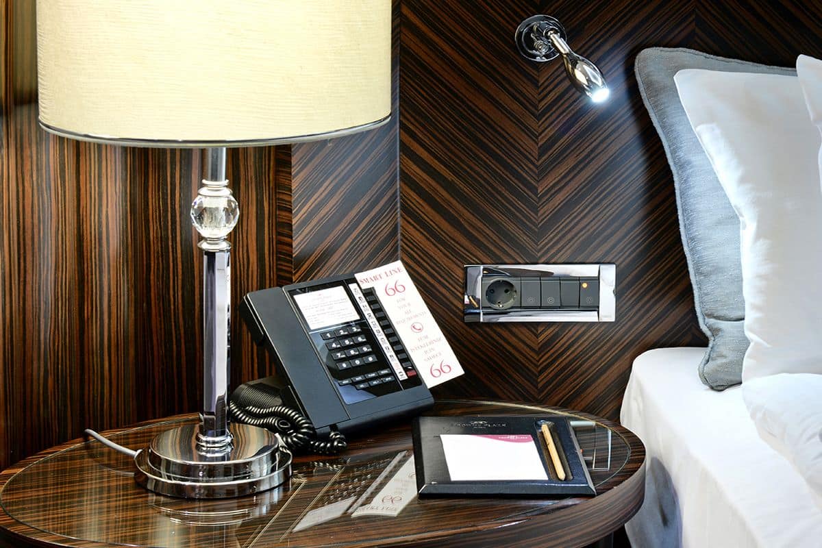 A hotel room with a wooden wall, a bedside table with a lamp, a telephone, and a bed with pillows.