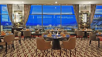 Un restaurante con varias mesas y sillas, amplias ventanas con vistas a la ciudad y alfombra estampada.
