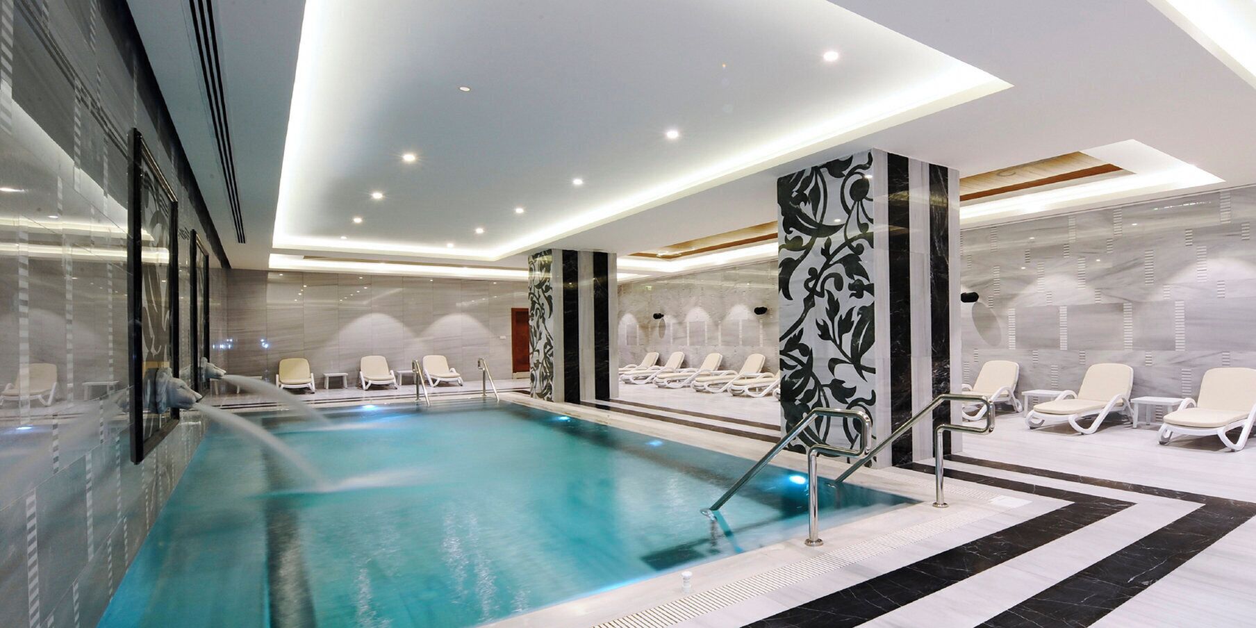 Una piscina interior moderna con sillones de descanso y una pared de azulejos de mármol.