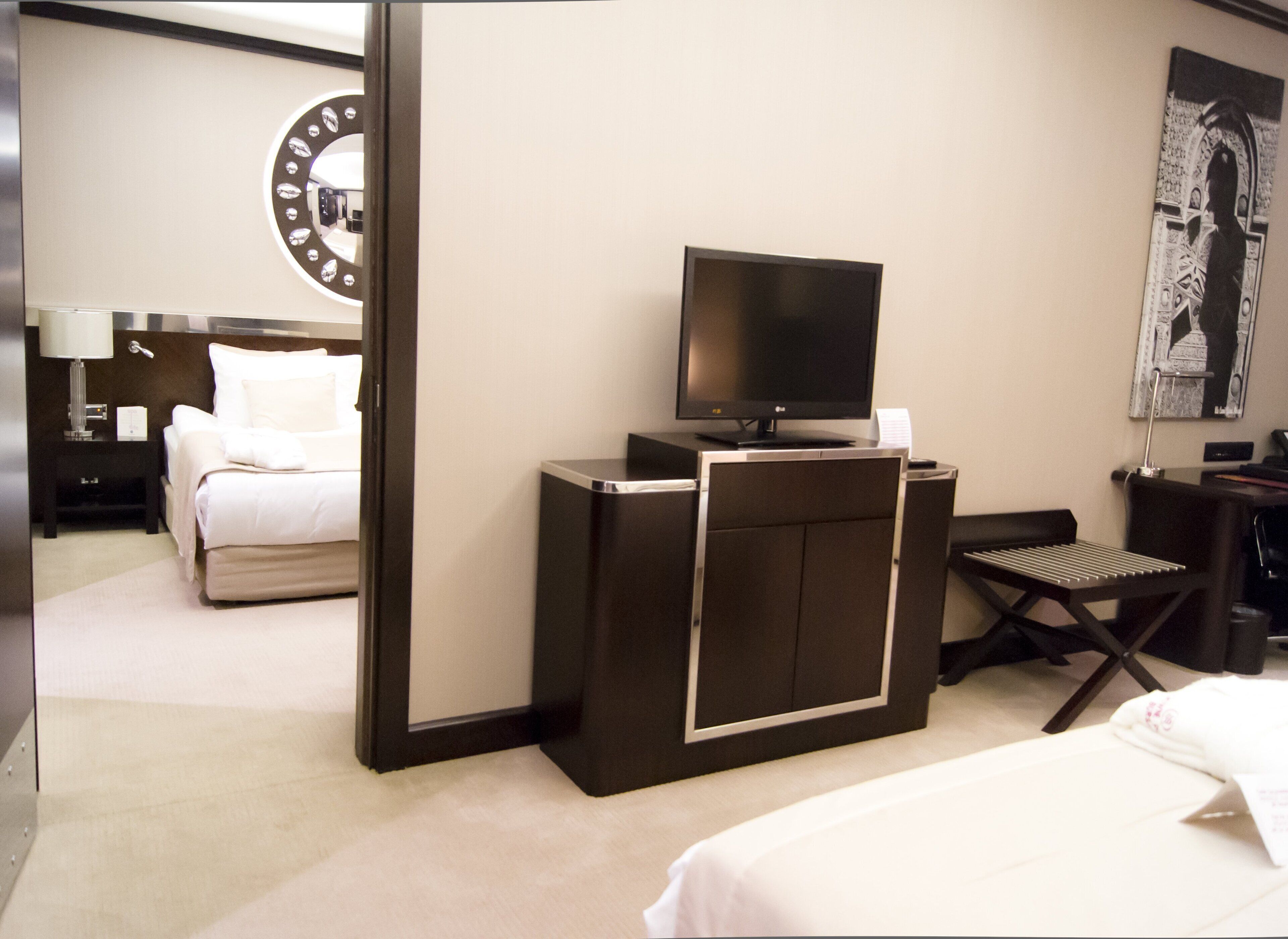 A hotel room with a bed, a desk with a flat-screen TV, a chair, a desk lamp, and a wall-mounted clock.