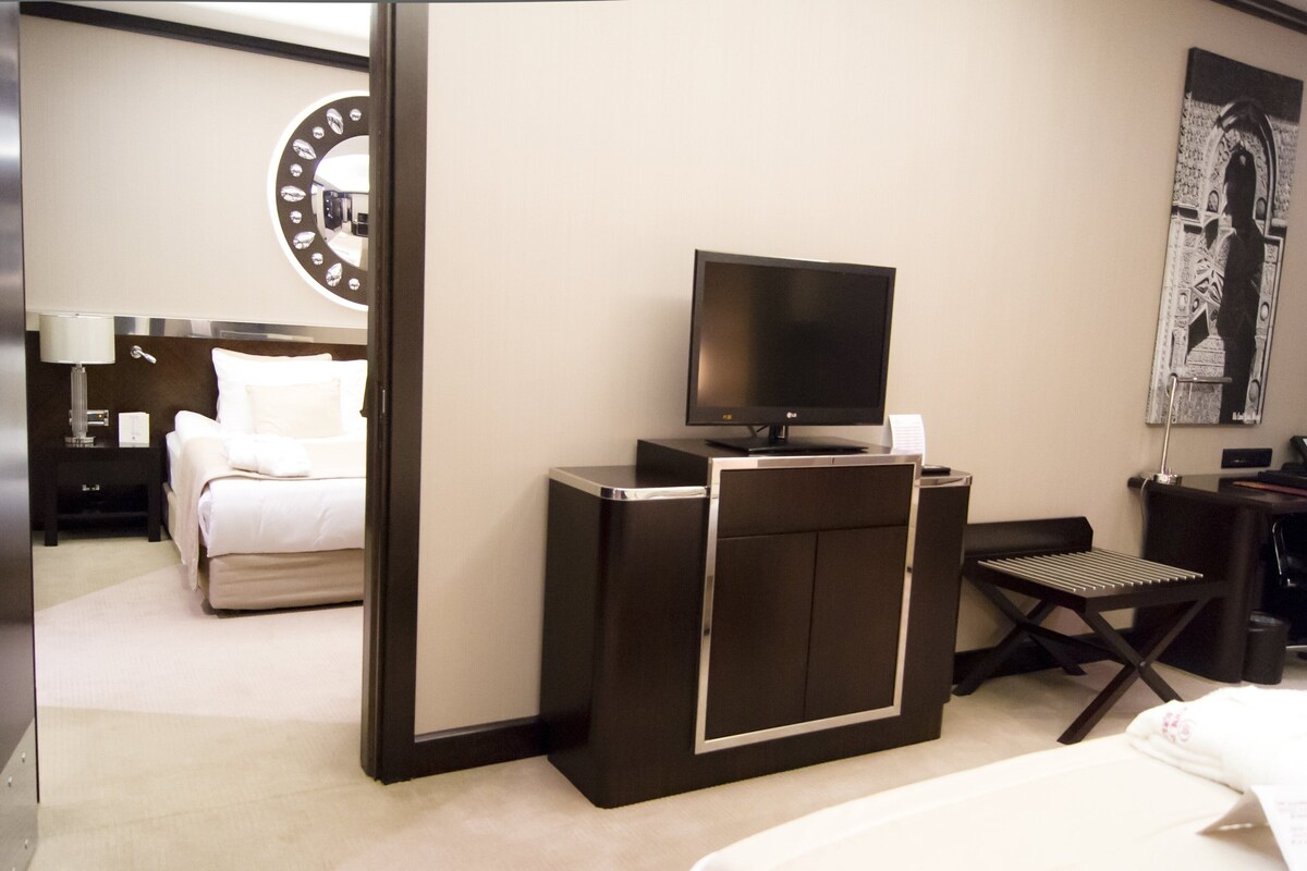 A hotel room with a bed, a desk with a flat-screen TV, a chair, a desk lamp, and a wall-mounted clock.