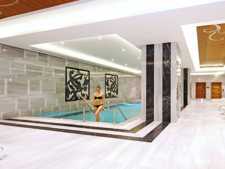 Un área de piscina interior moderna con una mujer en traje de baño, sillas de descanso y suelo de mármol.