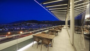 Zona de comedor en la azotea con mesas y sillas, con vistas a un paisaje urbano nocturno.
