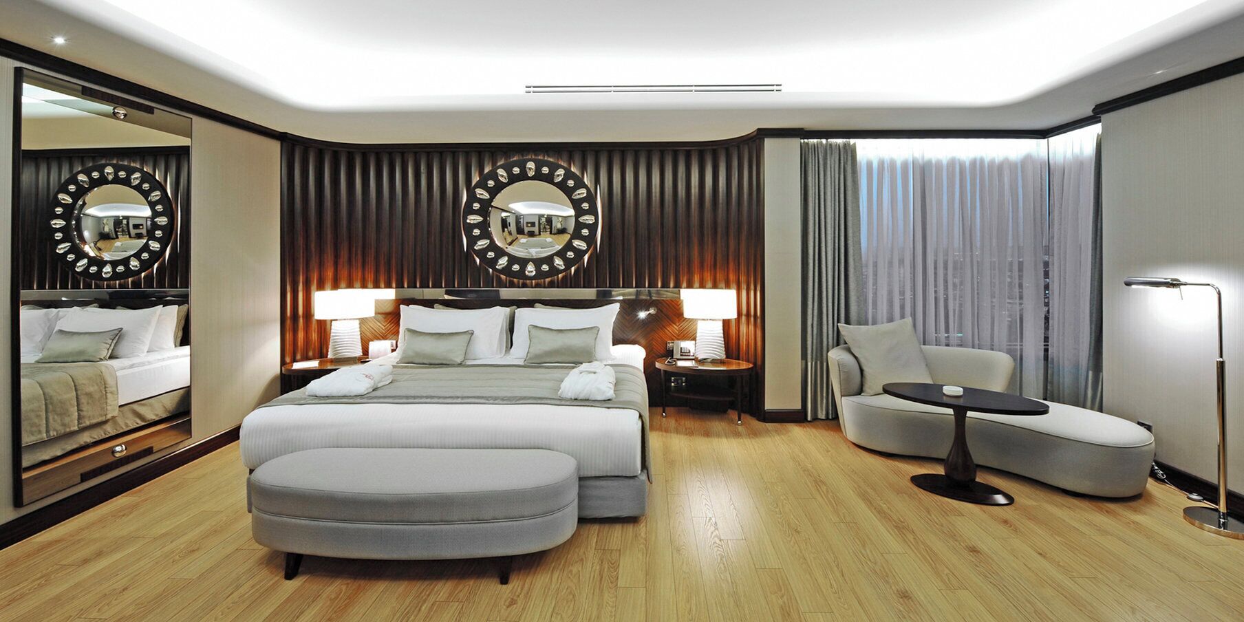 A modern hotel room with a large bed, two bedside tables with lamps, a sofa, a round table, and a large mirror with a circular frame.