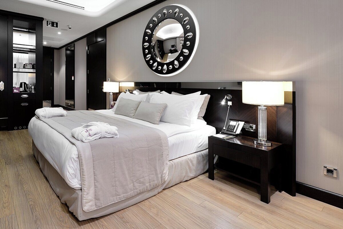A hotel room with a bed, bedside tables, a desk, a chair, a mirror, and a wardrobe.